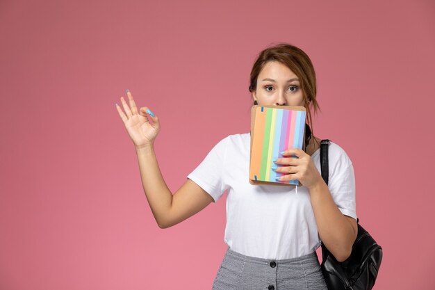 Jovem aluna de camiseta branca segurando cadernos com expressão de surpresa no fundo rosa.