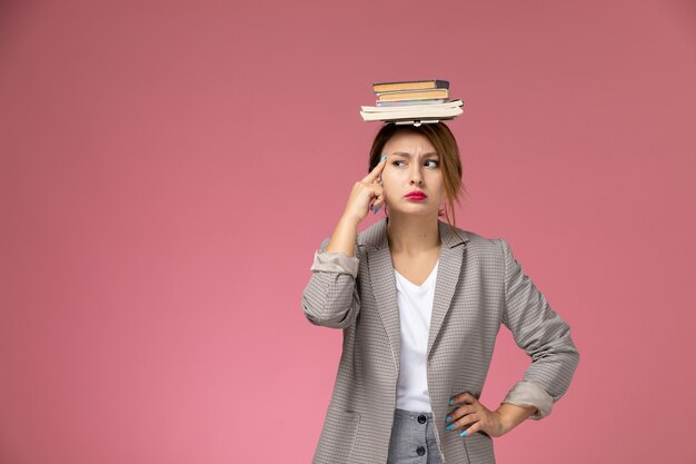 Jovem aluna de casaco cinza com cadernos na cabeça e expressão pensativa no fundo rosa lições faculdade estudo de frente