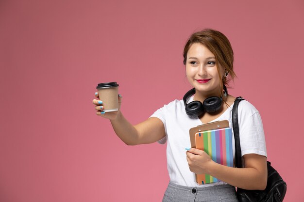Jovem aluna em camiseta branca com bolsa e fones de ouvido posando e sorrindo segurando café no fundo rosa lição