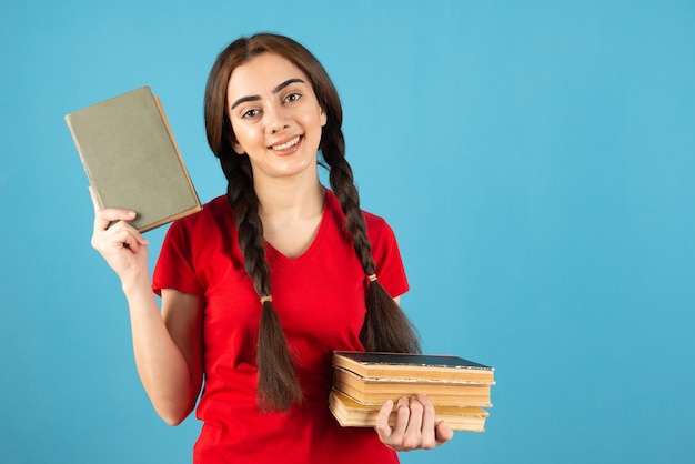 Jovem aluna em t-shirt vermelha com livros em pé na parede azul.