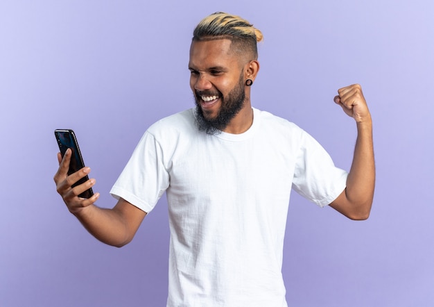 Foto grátis jovem americano africano com uma camiseta branca segurando o punho cerrado de smartphone feliz e animado, regozijando-se com seu sucesso em pé sobre um fundo azul