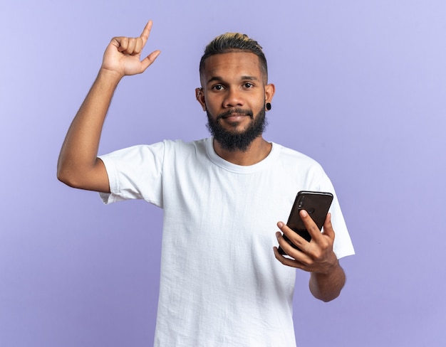 Jovem americano africano com uma camiseta branca segurando um smartphone, mostrando o dedo indicador olhando para a câmera, feliz e confiante novo conceito de ideia