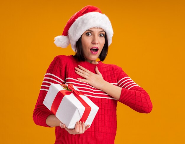 Jovem animada com chapéu de Papai Noel segurando um pacote de presente, olhando para a câmera, mantendo a mão no peito isolado em fundo laranja