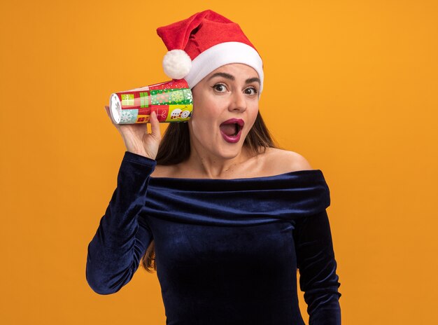 Jovem animada com um vestido azul e um chapéu de Natal segurando um copo de Natal, mostrando um gesto de escuta isolado em um fundo laranja