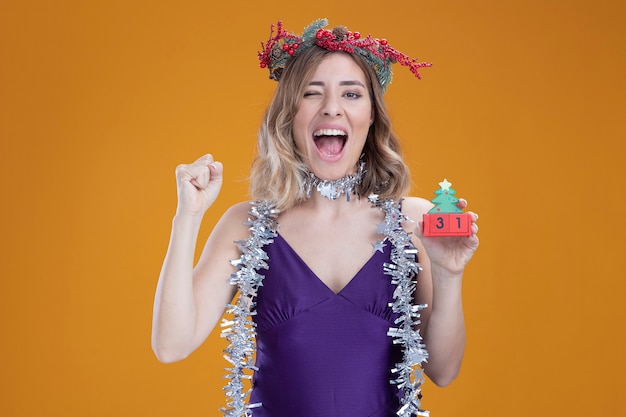 Jovem animada linda com vestido roxo e grinalda com guirlanda no pescoço segurando um brinquedo de natal mostrando um gesto de sim isolado em um fundo marrom