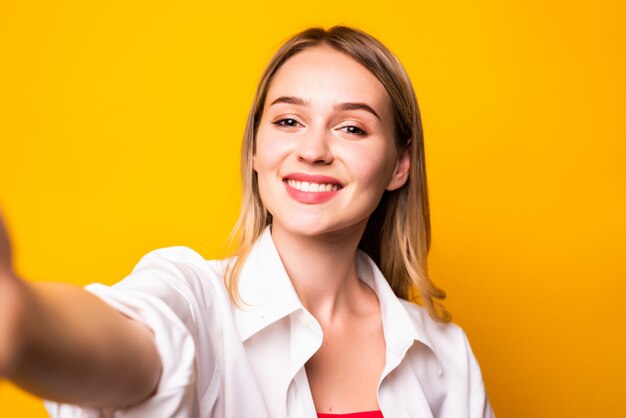 Jovem animada vestindo roupas casuais, isolada na parede amarela, tirando uma selfie com a mão estendida