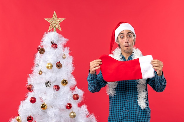 Jovem animado com chapéu de Papai Noel em uma camisa azul listrada e segurando uma meia de natal perto da árvore de natal