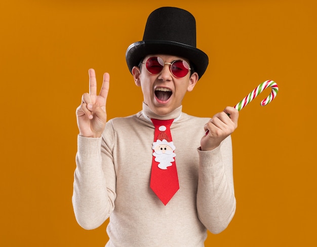 Jovem animado usando chapéu com gravata de natal e óculos segurando um doce de natal, mostrando um gesto de paz isolado na parede amarela