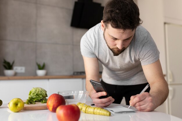 Jovem anotando a receita do smartphone na cozinha