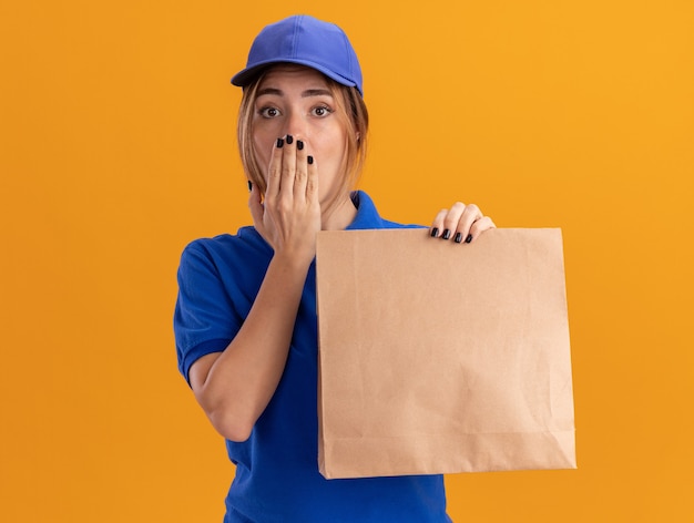 Jovem ansiosa, bonita, entregadora de uniforme, coloca a mão na boca e segura um pacote de papel em laranja