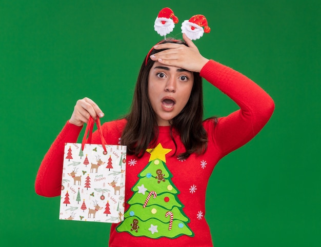 Jovem ansiosa caucasiana com bandana de Papai Noel coloca a mão na testa e segura uma sacola de papel isolada em um fundo verde com espaço de cópia