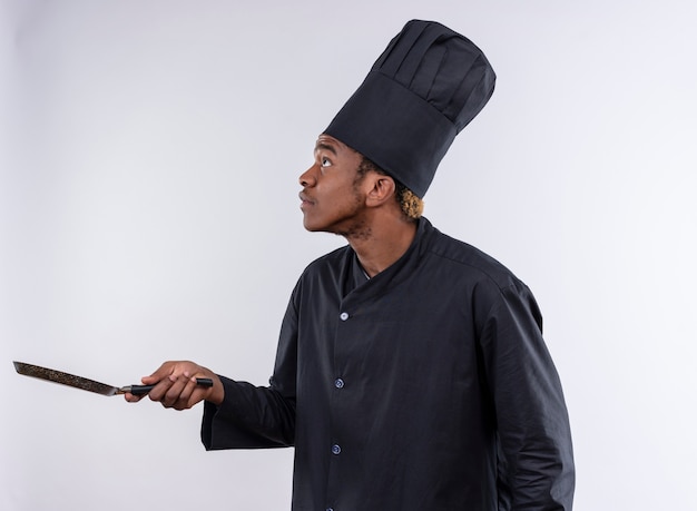 Jovem ansiosa cozinheira afro-americana com uniforme de chef fica de lado e segura a frigideira isolada no fundo branco com espaço de cópia