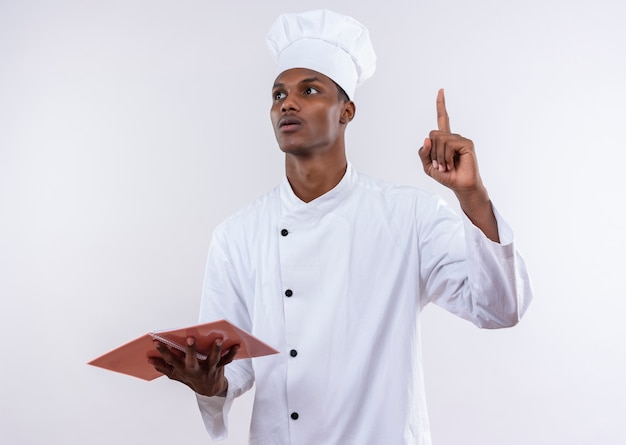 Jovem ansiosa cozinheira afro-americana com uniforme de chef segura um caderno e aponta, isolado no fundo branco com espaço de cópia