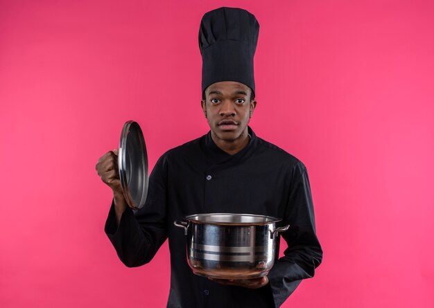 Jovem ansiosa cozinheira afro-americana com uniforme de chef segura uma panela e olha para a câmera isolada em um fundo rosa com espaço de cópia