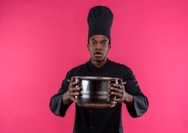 Jovem ansiosa cozinheira afro-americana com uniforme de chef segurando uma panela isolada em um fundo rosa com espaço de cópia