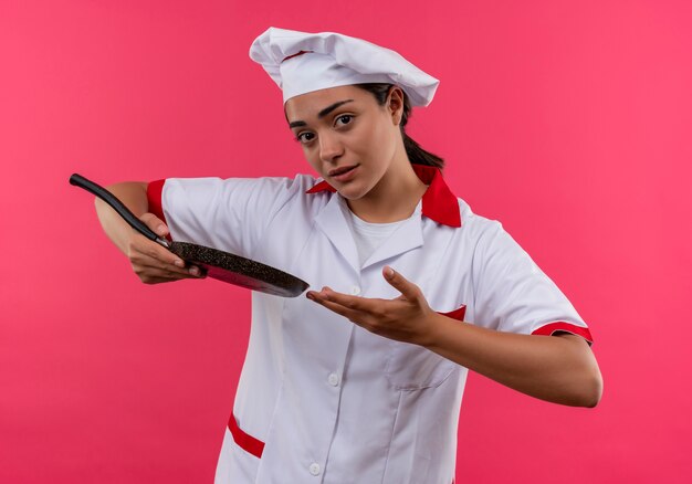 Jovem ansiosa cozinheira caucasiana com uniforme de chef segura a frigideira e aponta com a mão isolada em um fundo rosa com espaço de cópia