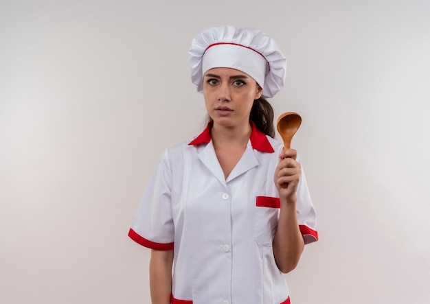 Jovem ansiosa cozinheira caucasiana com uniforme de chef segurando uma colher de pau isolada no fundo branco com espaço de cópia