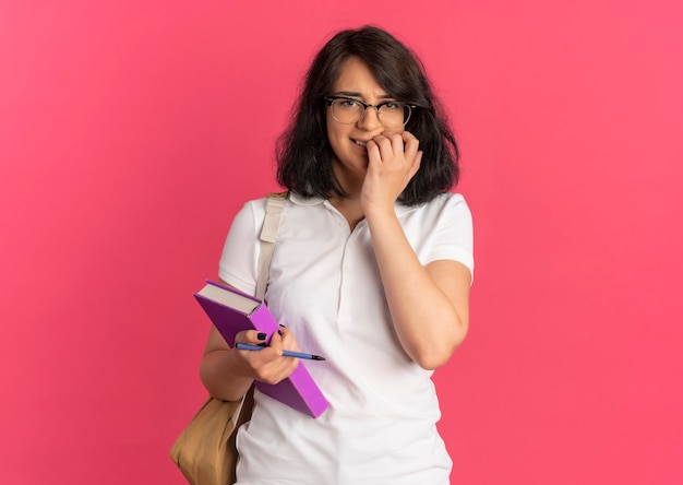 Jovem ansiosa e bonita caucasiana colegial usando óculos e bolsa nas costas morde os dedos segurando uma caneta e um livro olhando para a câmera no rosa com espaço de cópia