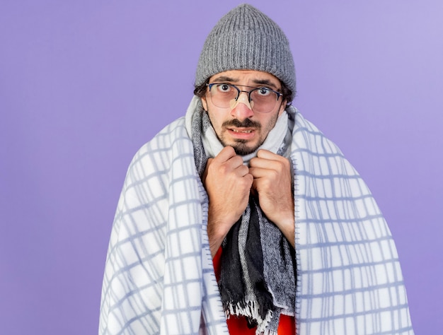 Foto grátis jovem ansioso, caucasiano, doente, usando óculos, chapéu de inverno e lenço embrulhado em xadrez, mantendo as mãos no peito, isolado na parede roxa com espaço de cópia