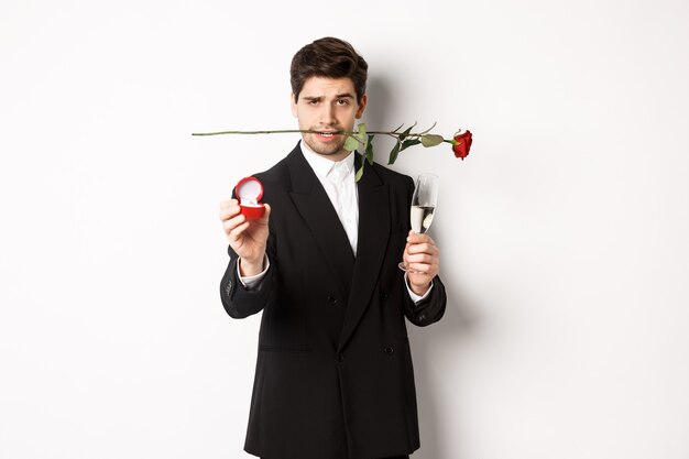 Jovem apaixonado de terno fazendo uma proposta, segurando uma rosa nos dentes e uma taça de champanhe, mostrando o anel de noivado, pedindo em casamento, em pé contra um fundo branco