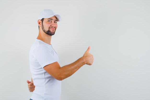 Jovem aparecendo o polegar de lado em uma camiseta branca, boné e parecendo feliz. vista frontal. espaço para texto