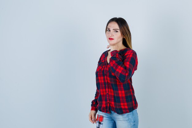 Jovem, apoiando o queixo na mão, em pé na pose de pensamento em camisa quadrada, jeans e olhando pensativa, vista frontal.