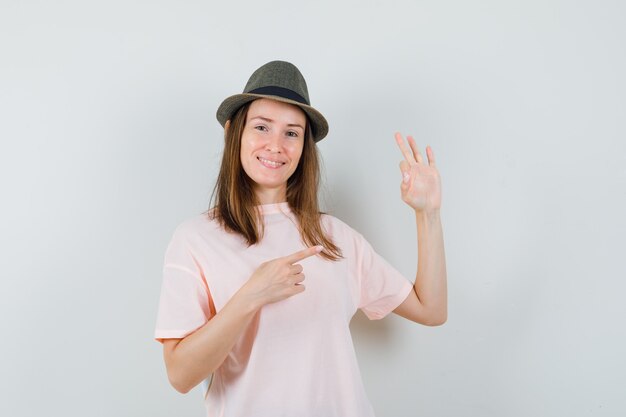 Jovem apontando de lado, mostrando o gesto ok em camiseta rosa, chapéu e parecendo confiante, vista frontal.