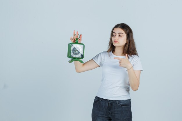 Jovem, apontando o relógio em t-shirt, jeans e olhando confiante, vista frontal.