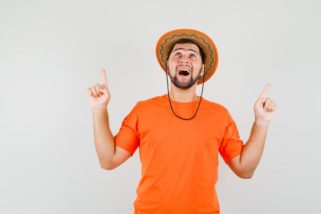 Jovem apontando os dedos para cima em t-shirt laranja, chapéu e olhando feliz, vista frontal.