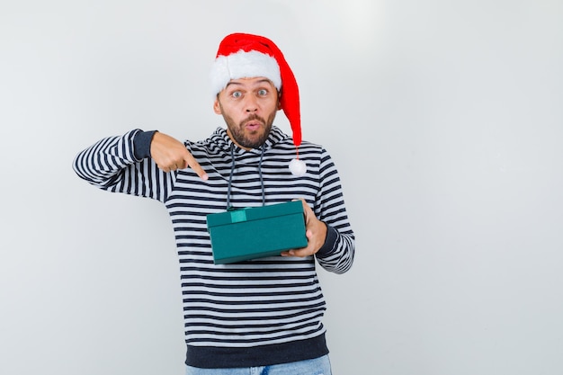 Jovem apontando para a caixa de presente com capuz, chapéu de Papai Noel e olhando maravilhado, vista frontal.