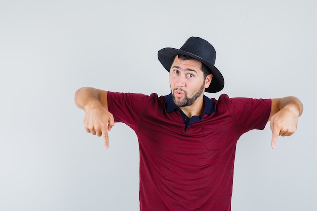 Jovem apontando para baixo em t-shirt vermelha, chapéu e parecendo surpreso, vista frontal.