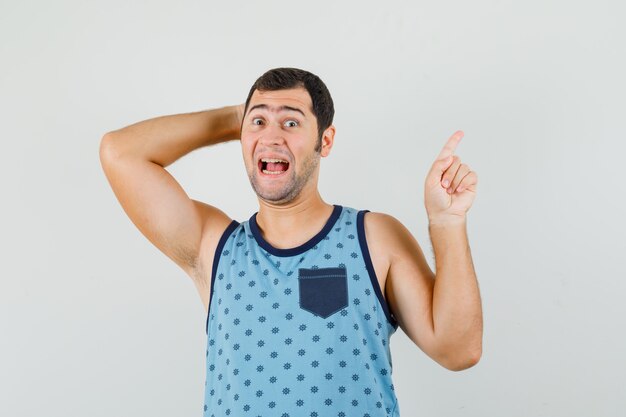 Jovem apontando para cima com a mão atrás da cabeça em camiseta azul e olhando alegre, vista frontal.