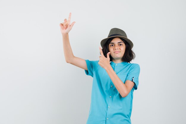 Jovem apontando para cima com camisa azul, chapéu e parecendo confiante.