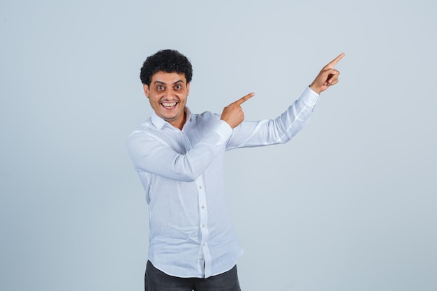 Jovem apontando para cima em camisa branca, calça e parecendo feliz. vista frontal.