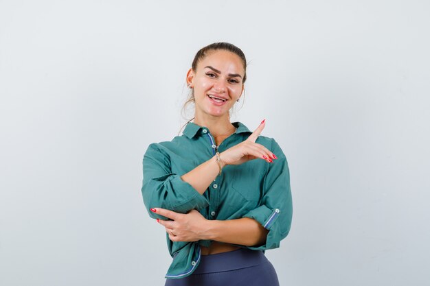 Jovem apontando para cima em camisa, calça e olhando alegre, vista frontal.