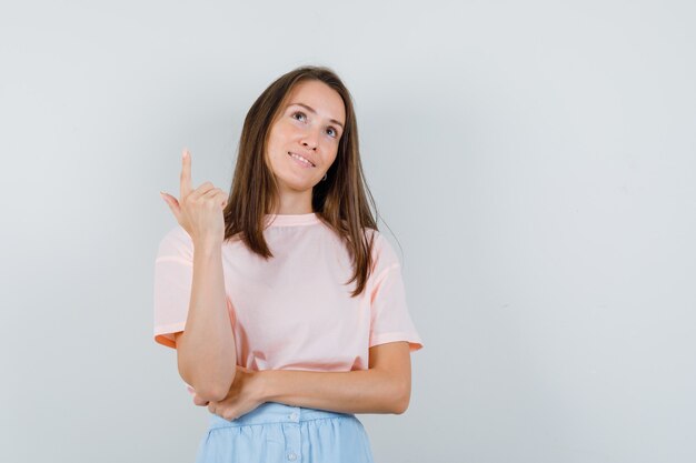 Jovem apontando para cima em camiseta, saia e olhando esperançosa. vista frontal.