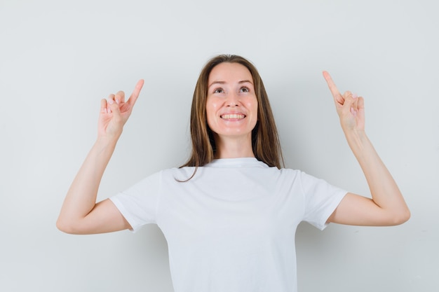 Jovem apontando para cima em uma camiseta branca e olhando alegre, vista frontal.