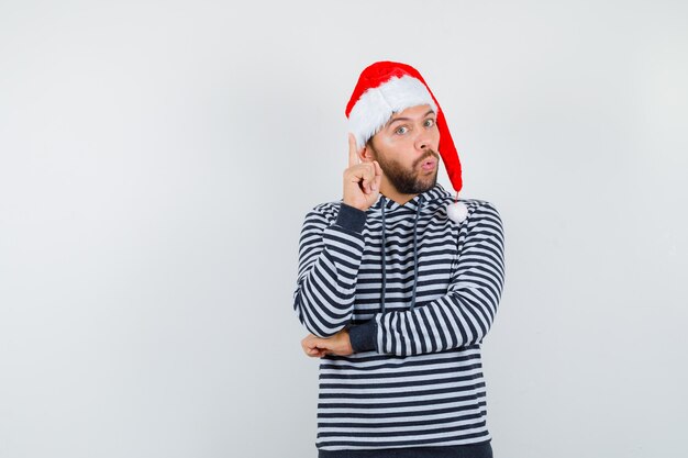 Jovem apontando para cima, fazendo beicinho com os lábios com capuz, chapéu de Papai Noel e parecendo chocado,