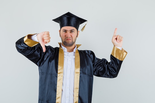 Jovem apontando para cima, mostrando o polegar para baixo em uniforme de pós-graduação e parecendo sombrio. vista frontal.