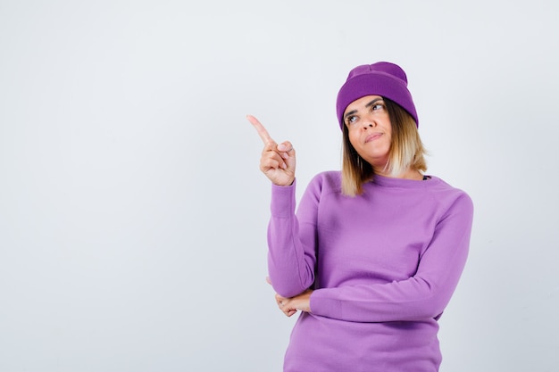 Jovem apontando para longe em um suéter roxo, gorro e parecendo satisfeito. vista frontal.