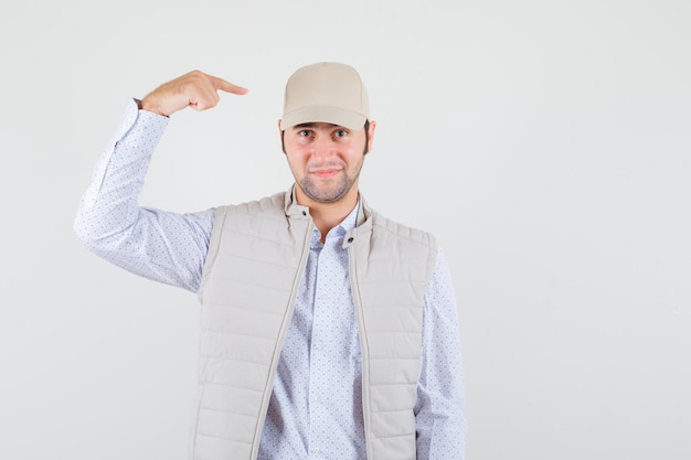 Jovem apontando para o boné de camisa, jaqueta sem mangas, boné e parecendo feliz. vista frontal.