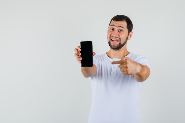 Jovem, apontando para o celular em uma camiseta branca e olhando feliz, vista frontal. espaço para texto