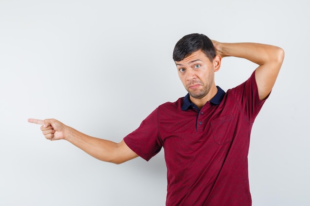 Jovem apontando para o lado com a mão atrás da cabeça em uma camiseta e parecendo hesitante. vista frontal.