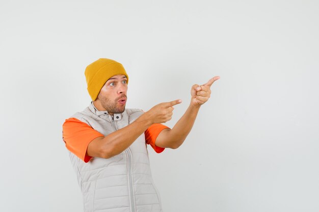 Jovem apontando para o lado com os dedos para cima na camiseta, jaqueta, chapéu e parecendo espantado.