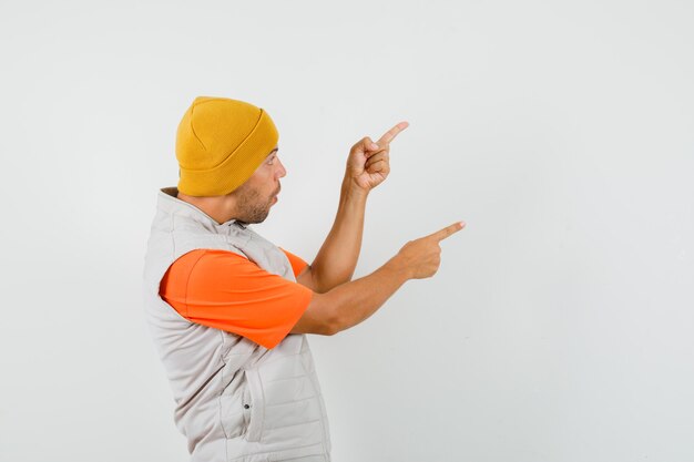 Jovem apontando para o lado com os dedos para cima na camiseta, jaqueta, chapéu e parecendo surpreso.