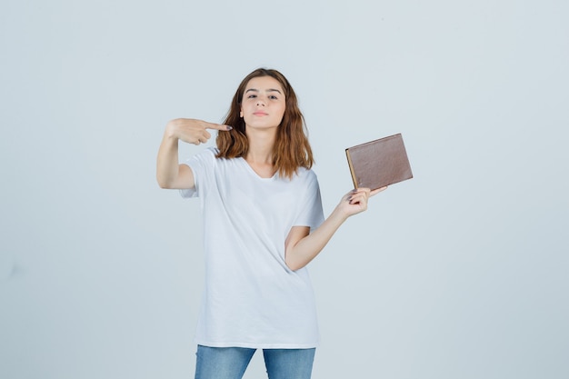 Jovem apontando para o lado direito enquanto segura o livro de camiseta, jeans e parece confiante. vista frontal.