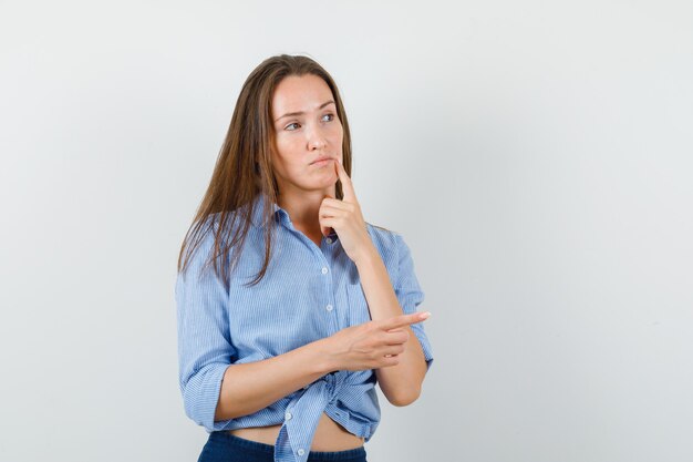 Jovem apontando para o lado em camisa azul, calça e olhando pensativa. vista frontal.