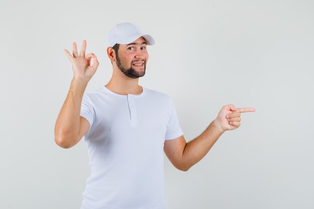 Jovem apontando para o lado enquanto mostra um gesto ok na camiseta, boné e parecendo satisfeito. vista frontal.