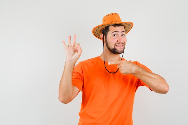 Jovem apontando para o seu sinal ok em t-shirt laranja, chapéu e parecendo satisfeito, vista frontal.