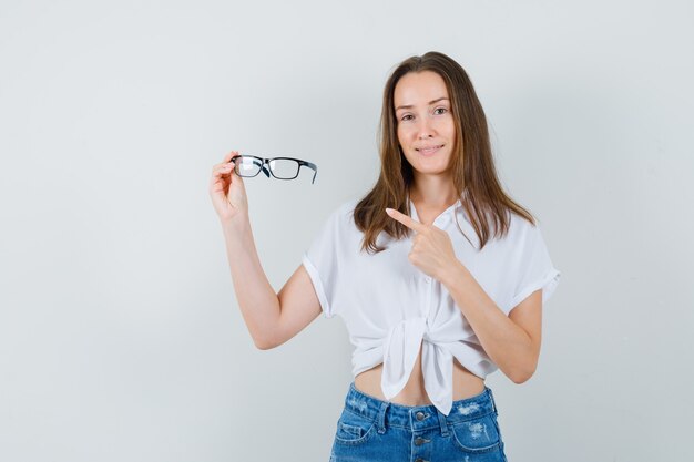 Jovem, apontando para os óculos na blusa branca e olhando positiva. vista frontal.